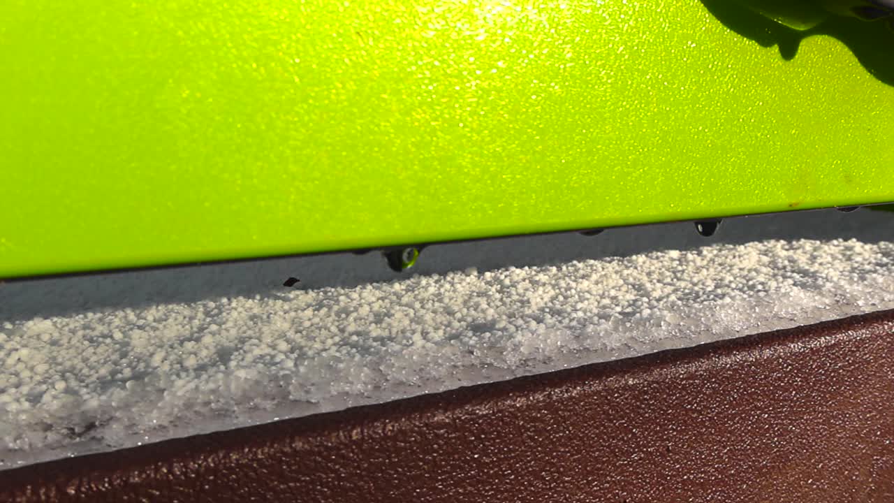 Close up view gliding in front of a playground wall where white fresh snow is melting and water drops are visible in the warm sunlight. Green and brown structure walls outside