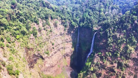 drone view of tad fane waterfall in pakse, laos, featuring twin falls dropping from a towering cliff into a deep, forest-covered gorge surrounded by vibrant greenery and mountainous terrain