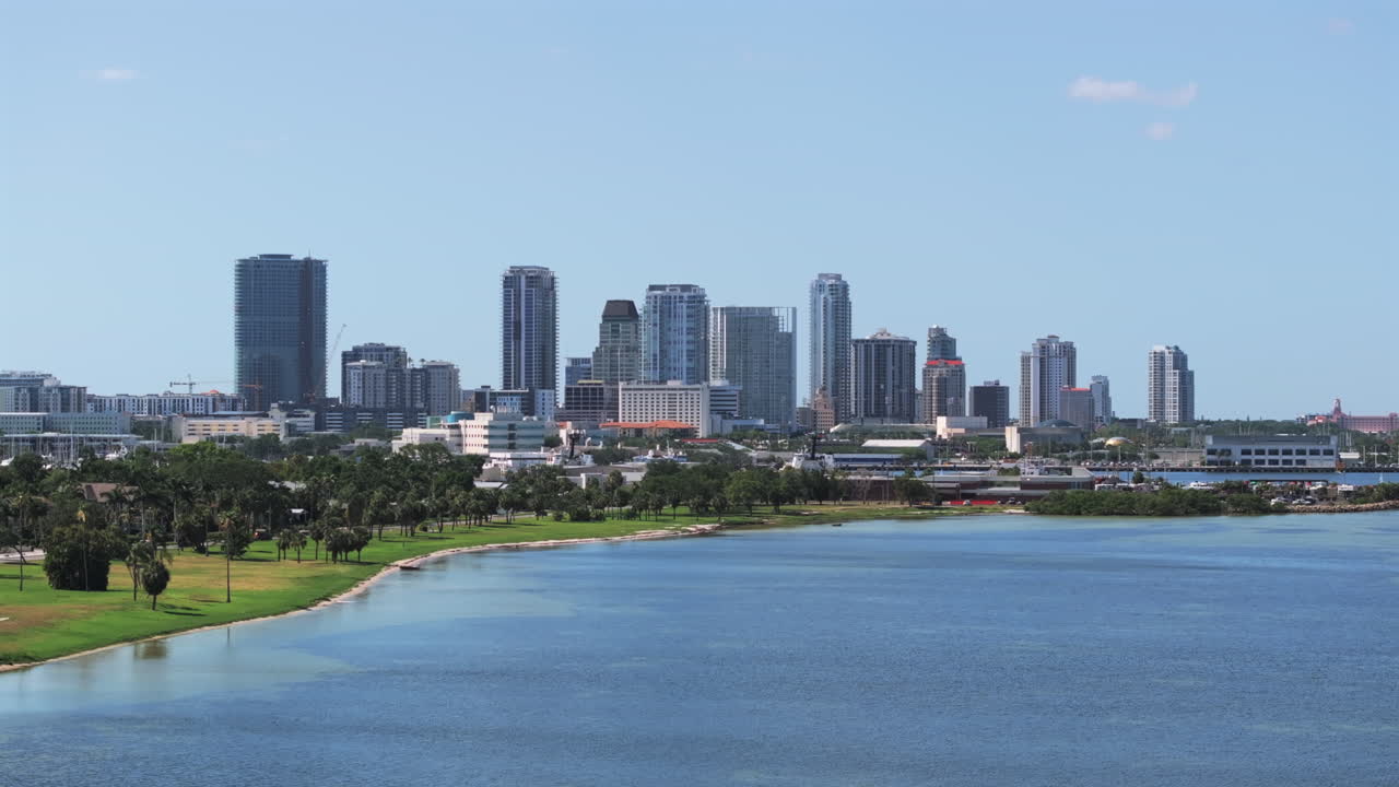 A sweeping aerial view of downtown St. Petersburg, Florida, featuring modern skyscrapers and condos, lush green Lassing Park,, and calm intrcoastal bay waters.