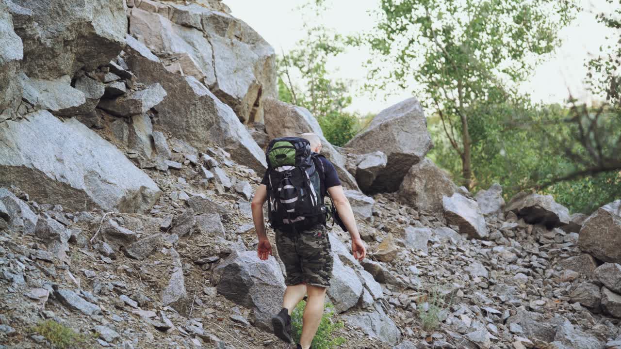A traveler with a backpack on his shoulders rises to the top of the mountain with stones in the afternoon in warm weather. Tourism.