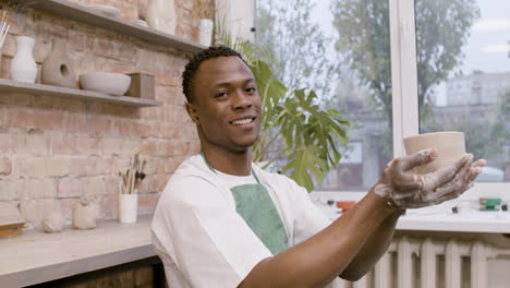 american clerk man holding at a ceramic piece that he has modeled on the potter wheel and looking at camera