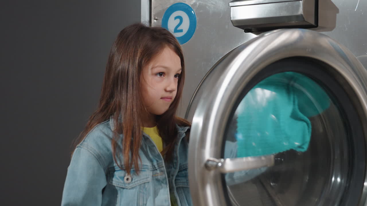 Zoom in on girl in denim jacket cleaning inner door of cloth washer inside laundromat, uses turquoise cloth through glass, label two visible on stainless machine, careful wiping motion