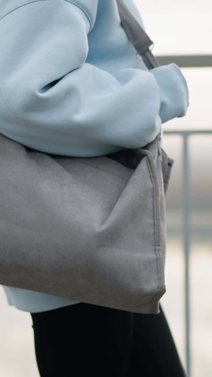 Partial view of young woman wearing blue sweater and black leggings, holding bag while walking calmly, background features iron railing and serene river view