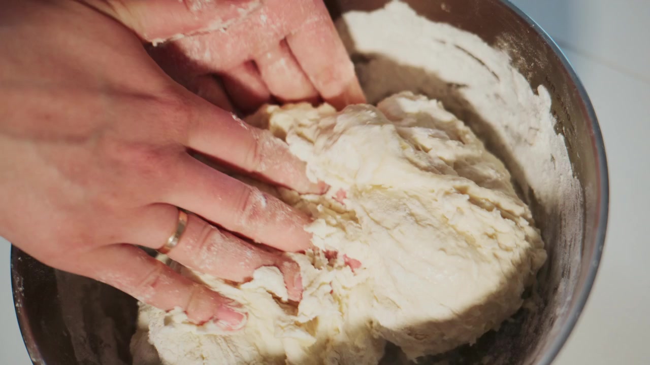 Hands knead dough in metal bowl. Flour dusts fingers and bowl surface. Person focused, calm, and skilled. Ideal for cooking, baking, or recipe videos. Shows traditional bread-making process