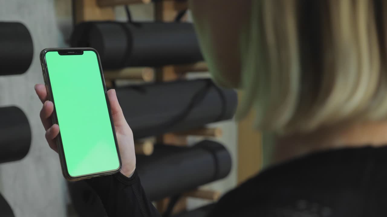 vista trasera de fitness mujer usando teléfono inteligente de pantalla verde en casa. mujer preparándose para el entrenamiento usando teléfono inteligente. mujer haciendo un toque en el teléfono móvil mientras descansa durante el entrenamiento de fitness