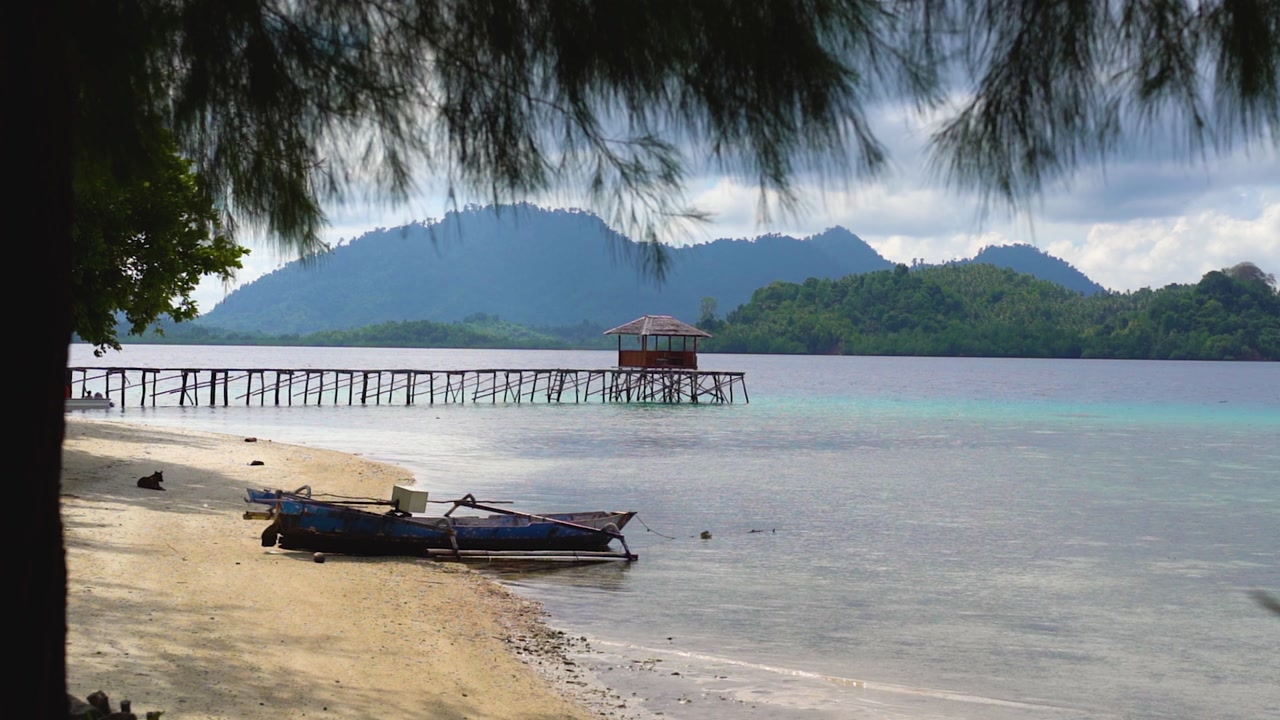 POV from a small beach hut on a tropical island in the Togean Islands in Sulawesi, Indonesia. The view is looking out at the beautiful beach with a jetty coming out from the island.