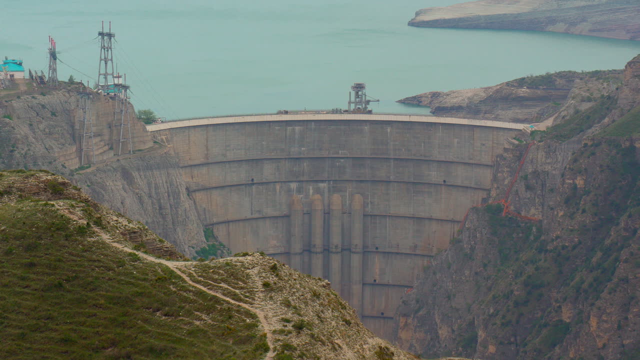 massive concrete arch dam between rocky canyon walls with calm reservoir water power lines engineering structures and dramatic mountain infrastructure landscape Telephoto video frame