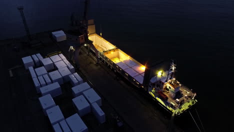 Crane-loading-shipping-containers-on-a-ferry-at-night---tilt-down-aerial-view