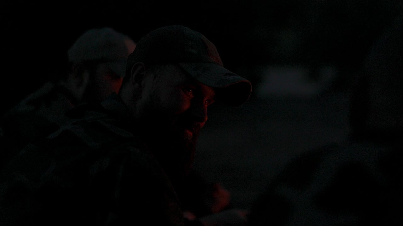 close-up no rosto de um homem com barba em roupas de camuflagem e boné à noite iluminado por chamas de fogueira, conta história assustadora para amigos, piadas, acampamento noturno, caça
