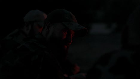primer plano en la cara de un hombre con barba en ropa de camuflaje y gorra por la noche iluminado por las llamas de la fogata, cuenta historias aterradoras a los amigos, bromas, campamento nocturno, caza