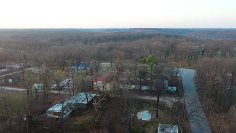 middle of nowhere houses surrounded by barren forest trees in tiny rural town with backroads on dark cloudy day - aerial drone pullback