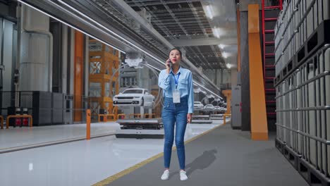 cuerpo lleno de una trabajadora profesional asiática de pie en la fábrica de automóviles ev, instalación industrial de alta tecnología, ella está hablando con alguien en el teléfono