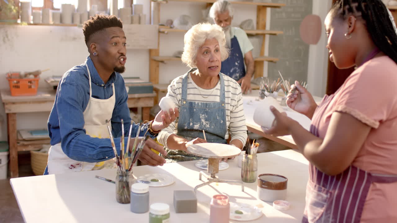 Happy diverse group of potters glazing clay jugs and discussing in pottery studio