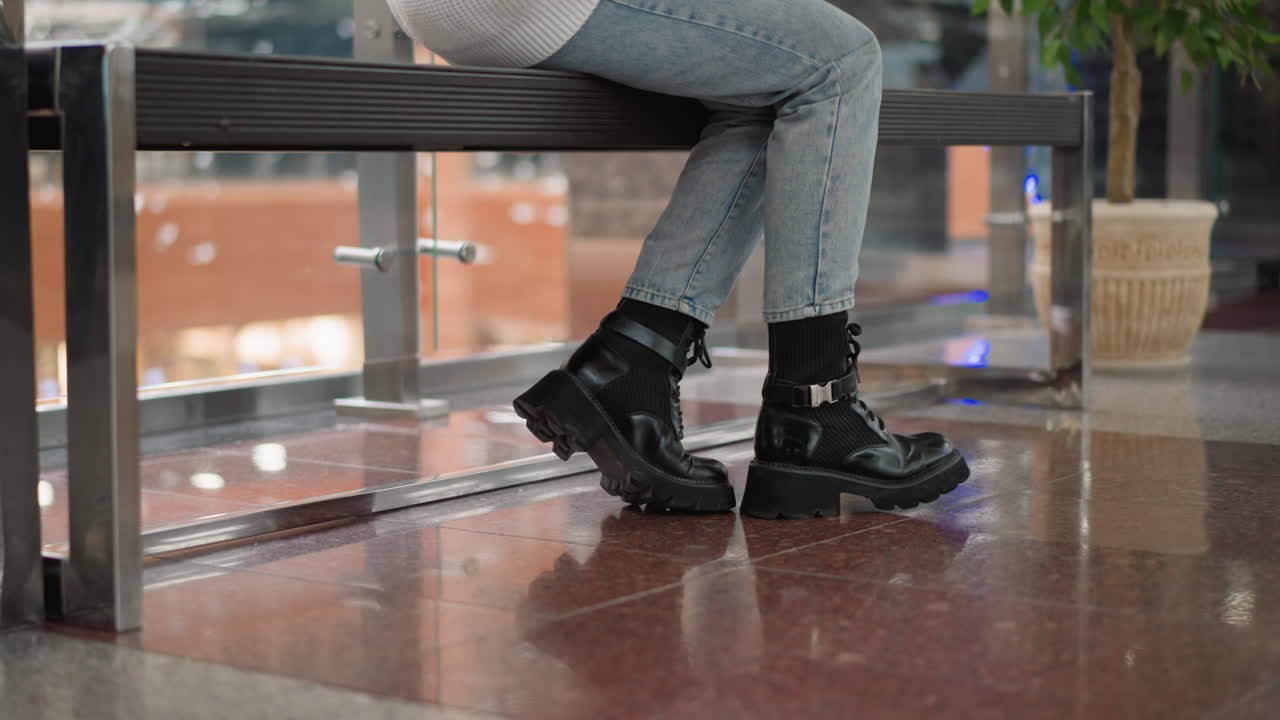 poised figure walking to seat carrying handbag, legs crossed midstride on polished mall floor, casual jeans and boots detail, ambient reflections and slow elegant motion toward bench