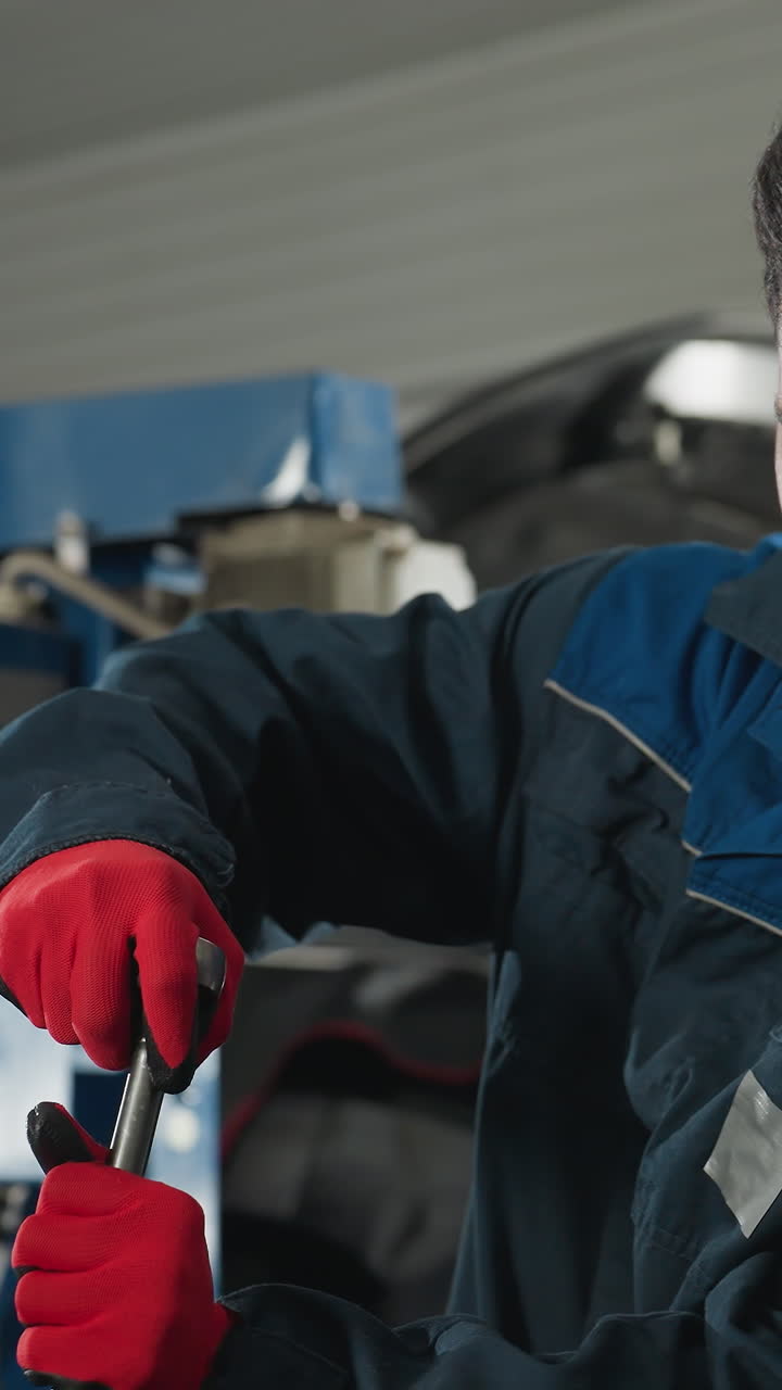 ingeniero automotriz con uniforme de trabajo azul y guantes rojos sonriendo mientras repara el motor del automóvil en el taller, el fondo presenta herramientas industriales, capó abierto del automóvil y equipos mecánicos