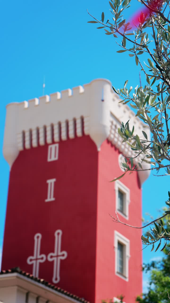 Green tree moving in the wind near the red tower of the Cremat Castle Winery over blue sky. Vertical