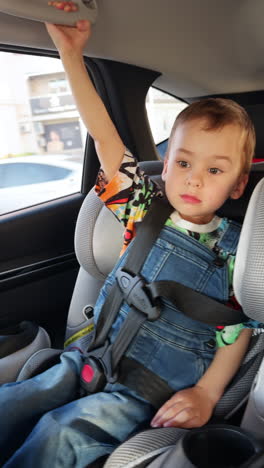 lovely caucasian kid wearing a t-shirt and jeans romper sits in baby car seat. child travelling in automobile. vertical video.