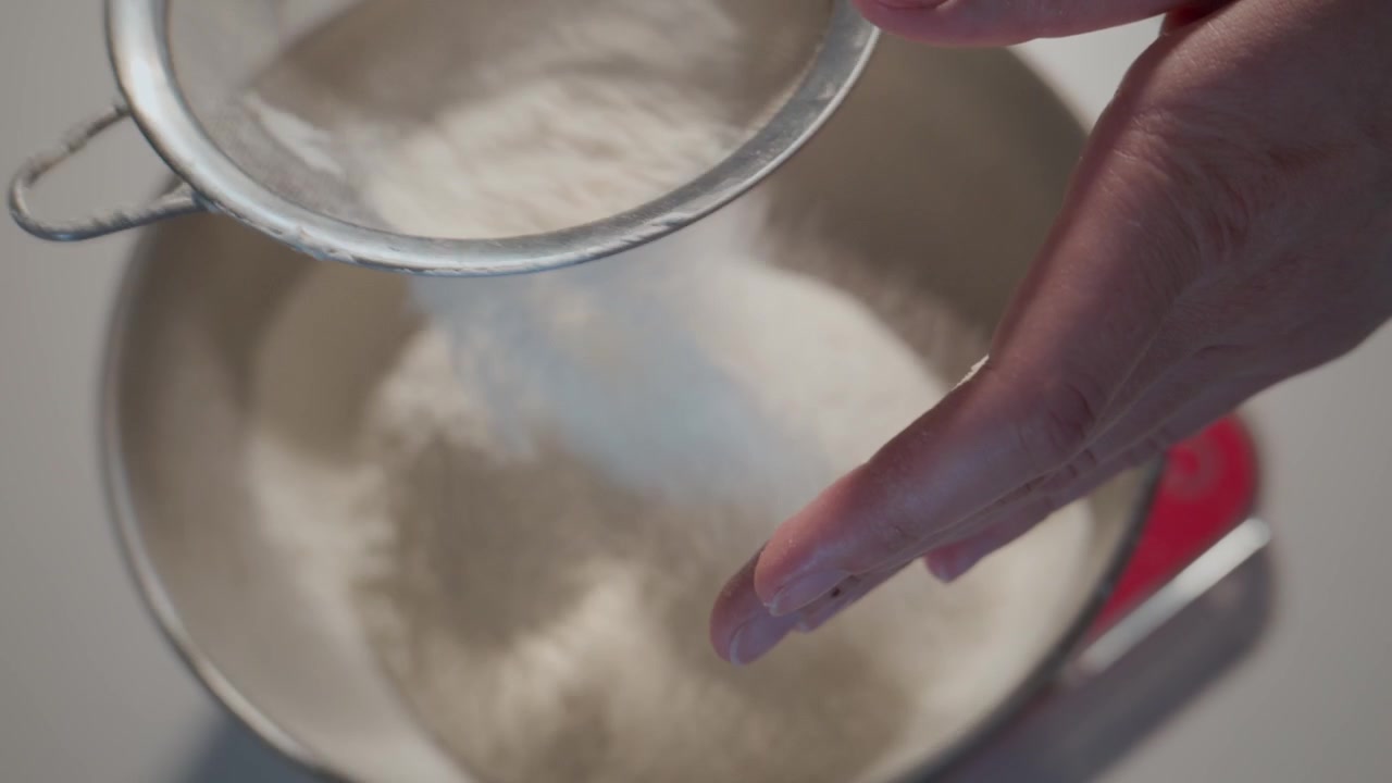 Una mano setaccia la farina in una ciotola. Un primo piano evidenzia la preparazione per la cottura. Ideale per ricette, tutorial di cucina o blog di cibo.