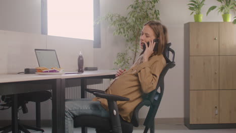Mujer-Embarazada-Hablando-Por-Su-Teléfono-Móvil-Mientras-Está-Sentada-En-El-Escritorio-Durante-El-Almuerzo-De-La-Oficina