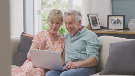 senior retired couple sitting on sofa at home making video call on laptop