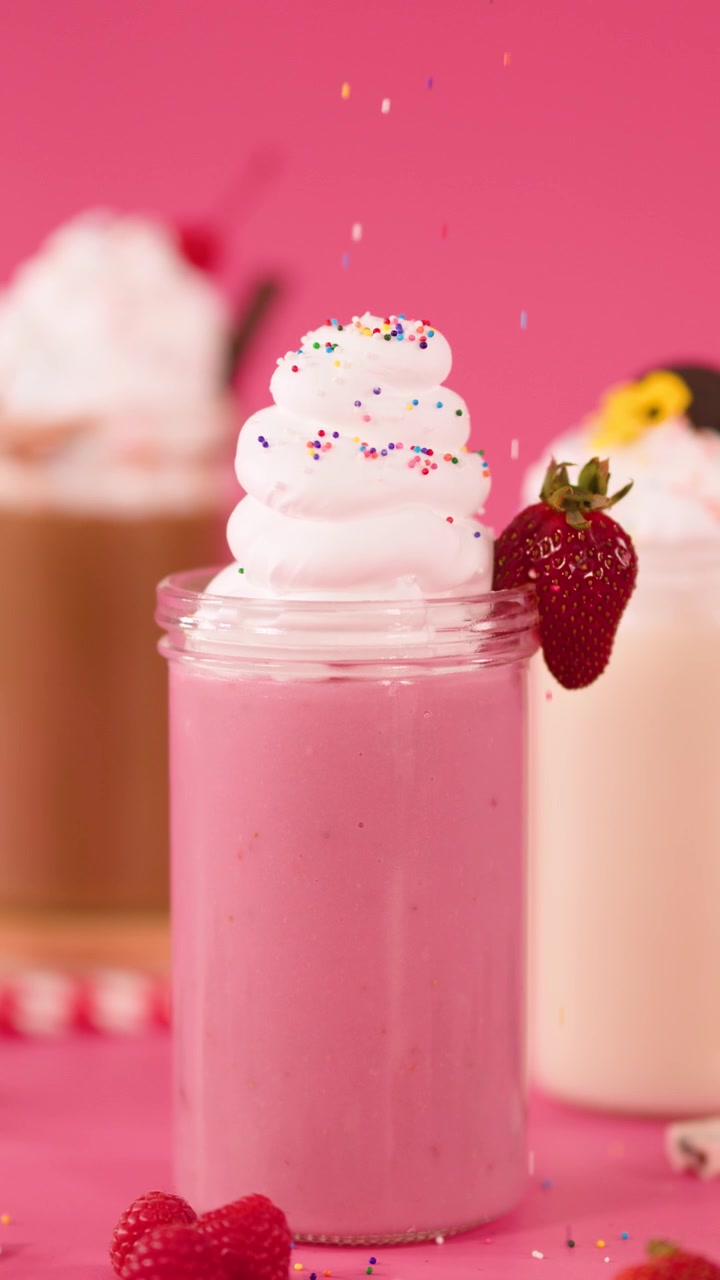 Frullato rosa alla fragola guarnito con panna montata, codette di zucchero e una ciliegia, sotto una luce brillante da studio.