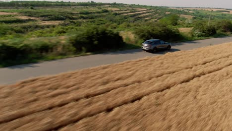disparo de seguimiento aéreo, de un coche deportivo suv de lujo gris, conduciendo por una carretera asfaltada de campo, rodeado de campos de trigo dorados