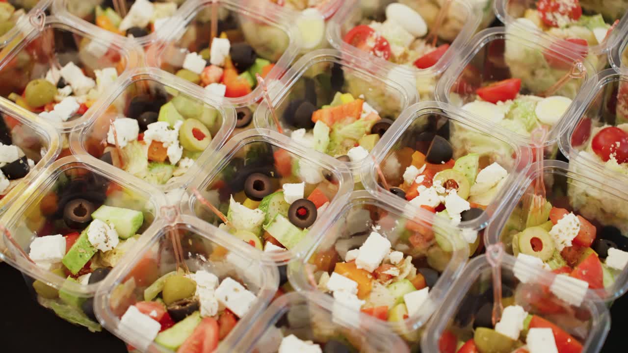 Dozens of individual grab-and-go containers are packed with fresh, healthy Greek salad with feta cheese and olives, ready for a catered event, delivery