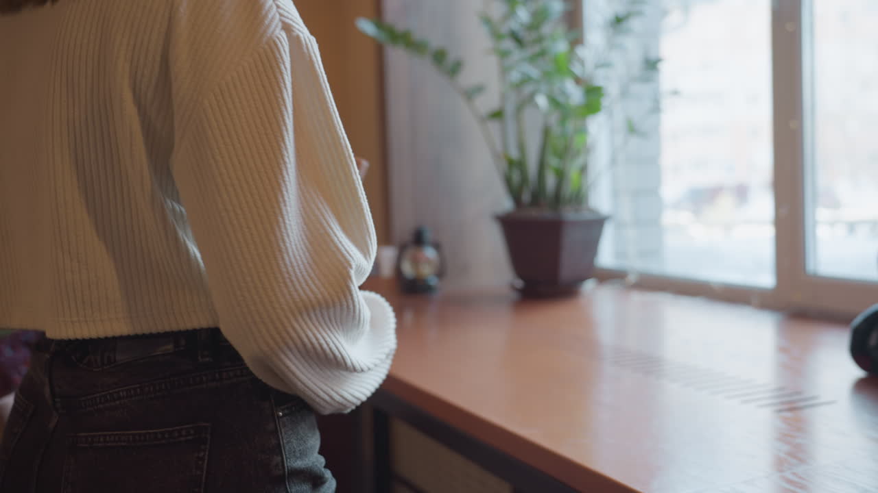 young woman in ribbed sweater sitting at table carefully adjusts book in hand with folded colorful cloth placed beside her under soft indoor lighting creating cozy atmosphere