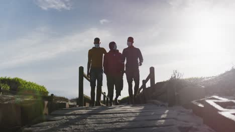 african american senior father and two teenage sons wearing face masks walking on the beach