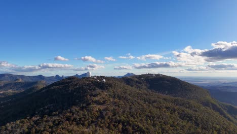 aerial drone footage glides over forested hills toward siding spring observatory under clear blue skies, revealing telescopes and expansive australian landscape in daylight