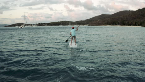 un dron sigue a un surfista de remo que rema hacia una isla montañosa en el caribe