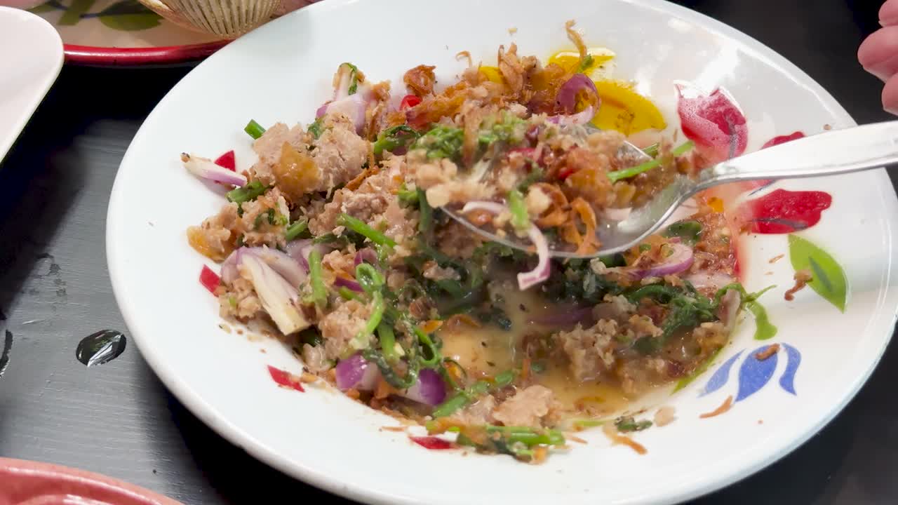 Hand uses spoon to mix spicy minced pork salad with herbs and vegetables in a brightly lit Thai restaurant setting, close-up overhead view