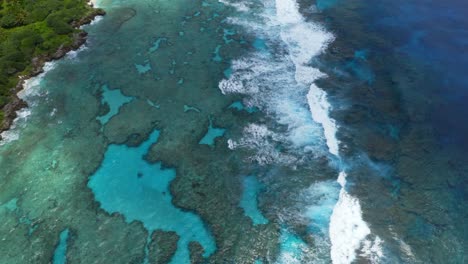 the drone's camera pans over the blue lagoon, capturing the stunning transparency of the water, allowing a view of the seabed and the rocks beneath, while the waves crash on the reef