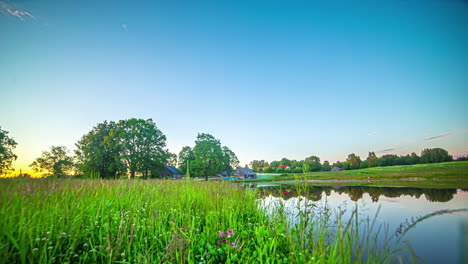 Timelapse-shot-of-few-cottages-beside-a-small-lake-surrounded-by-lush-green-vegetation-throughout-the-day