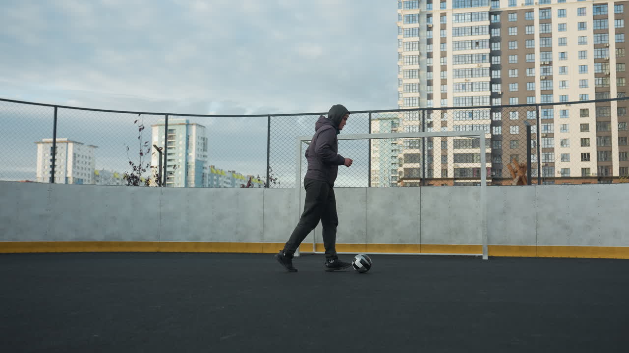 Man practicing soccer skills, controlling ball with precision while walking on outdoor sports ground, urban backdrop features high-rise buildings and goalpost