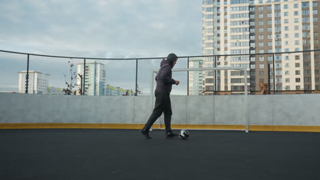 man practicing soccer skills, controlling ball with precision while walking on outdoor sports ground, urban backdrop features high-rise buildings and goalpost