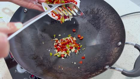 overhead view of hands adding chopped chili peppers and garlic into a heated wok on a stovetop, with bright natural lighting and minimal camera movement