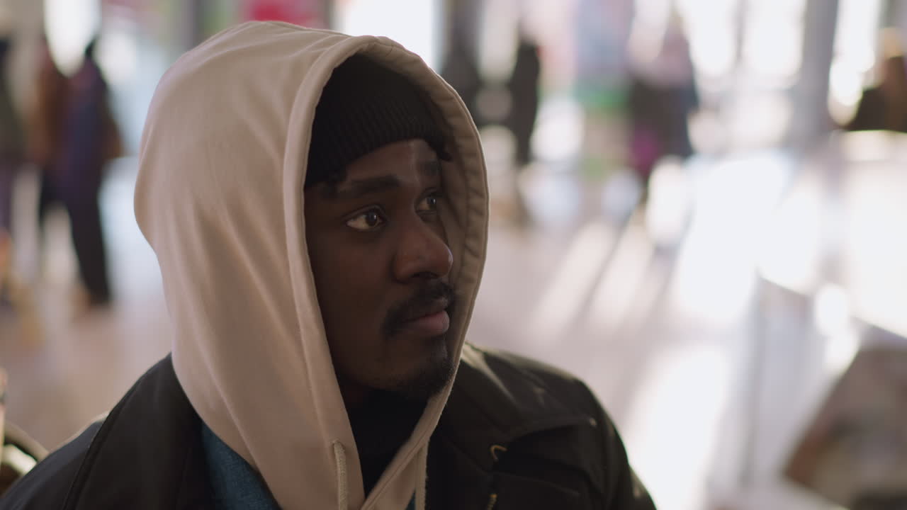 Close-up of young man with hoodie and beanie, looking upward with focused expression, soft background light and blurred people in modern shopping mall, side view
