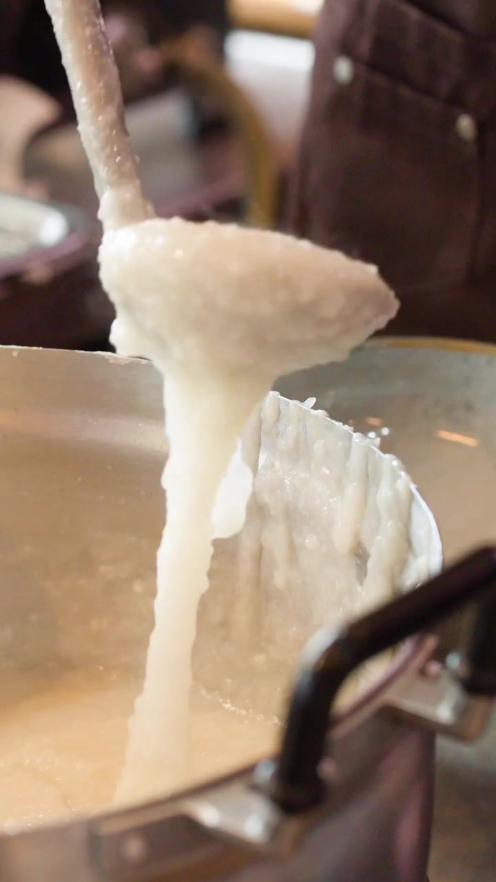 Close-up of ladle scooping thick white congee from a steaming metal pot in kitchen.
