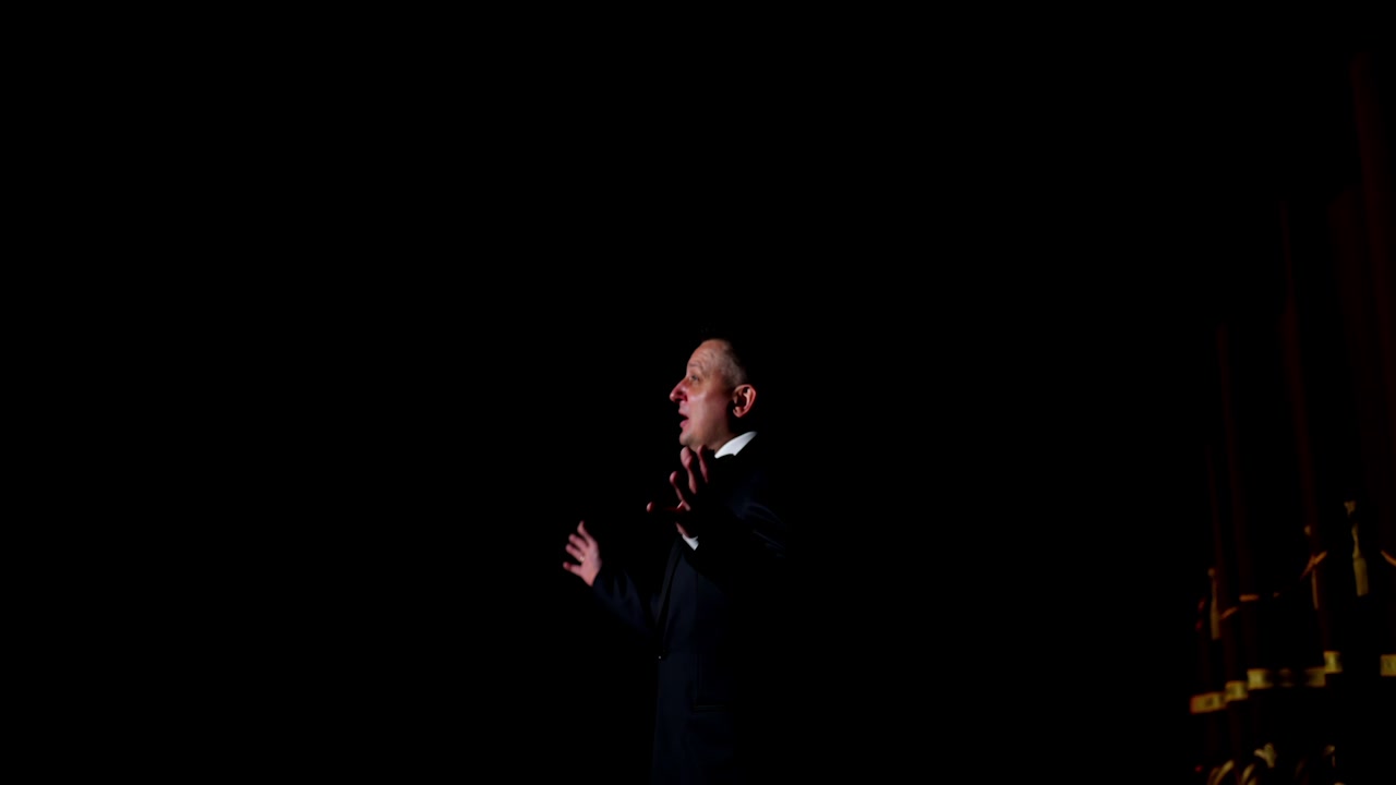 Speaker on black background. Profile view of a man talking to the audience while standing on stage. Performer in darkness.