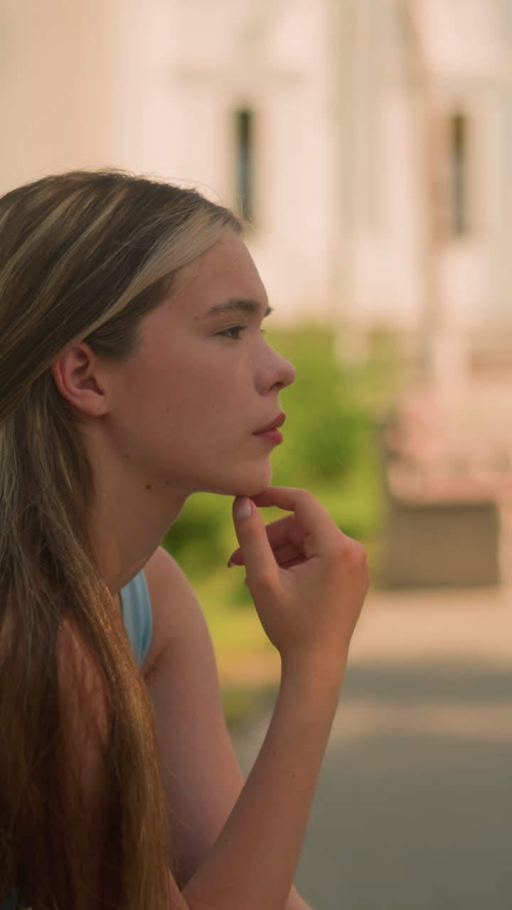 Young lady touching chin with left hand while seated outdoors, gazing thoughtfully into distance, background features building, greenery, and soft shadows under sunlight