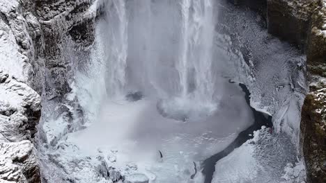 drone tilt up shot of powerful waterfall in iceland flowing between snow-covered cliffs and icy rocks. mist rises as water crashes into deep pool surrounded by rugged winter landscape
