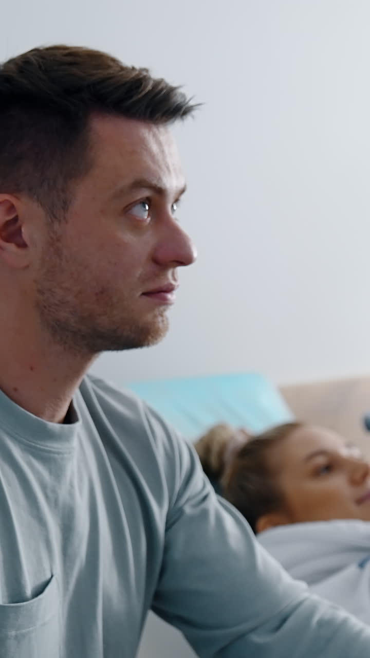 Focused bearded man looks up at the screen showing his future baby. Blonde woman lies on the couch at backdrop in blur. Vertical video