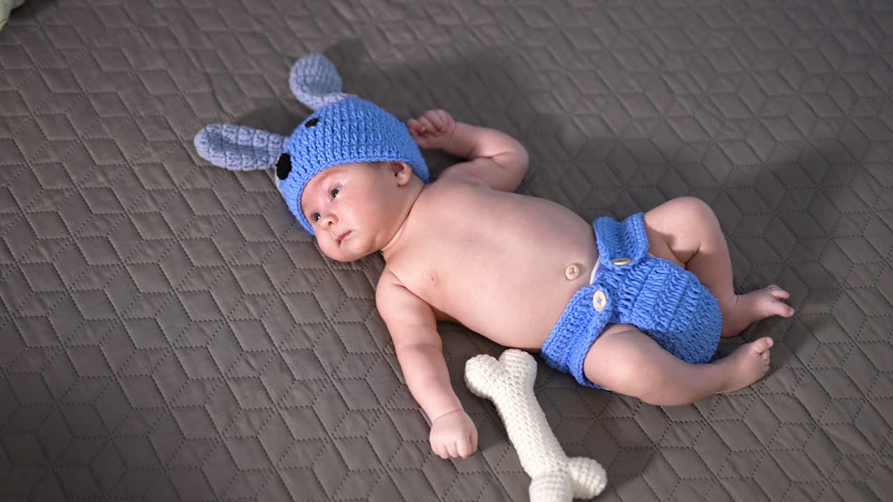 Cute baby in a funny blue suit lying on the bed. Puppy costume put on a newborn kid and funny soft knitted bone beside. View from above.