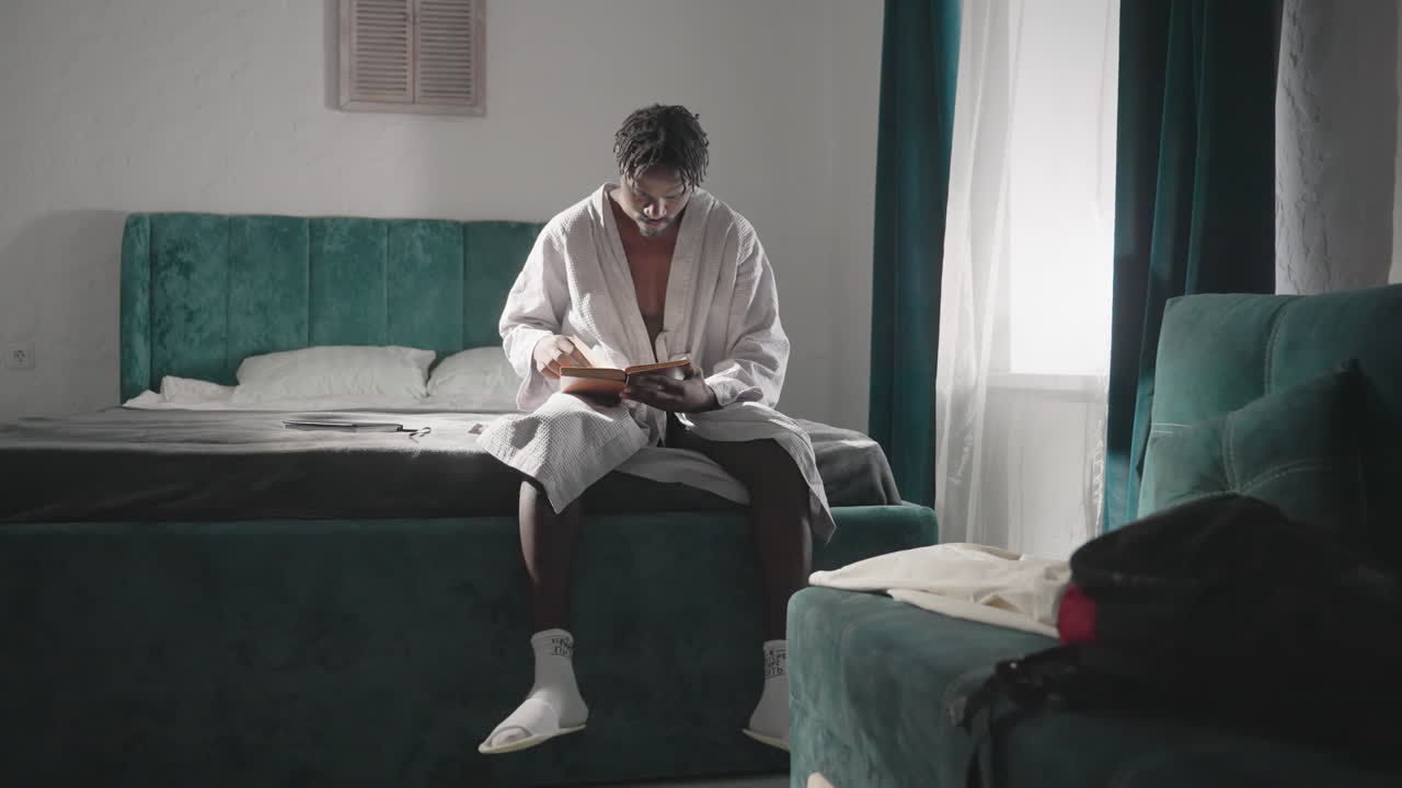 Man in white robe sitting on bed, reading large book with deep focus. Soft natural light flows through window, creating a peaceful, reflective mood in a cozy, serene indoor space