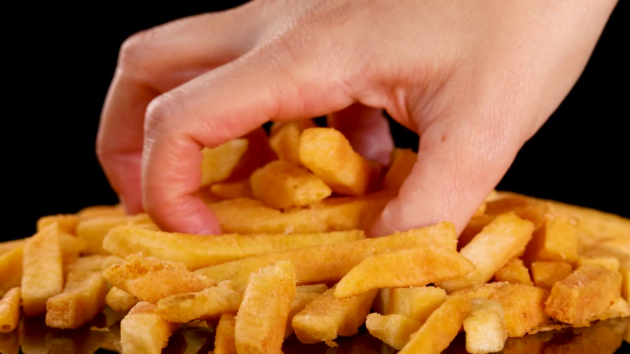 Hand grabs golden, crispy French fries from a pile under bright studio lighting, black background