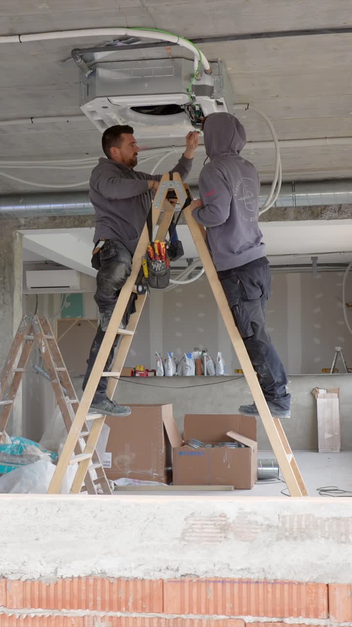 Two Men Installing Air Conditioning Unit on Ceiling at Construction Site