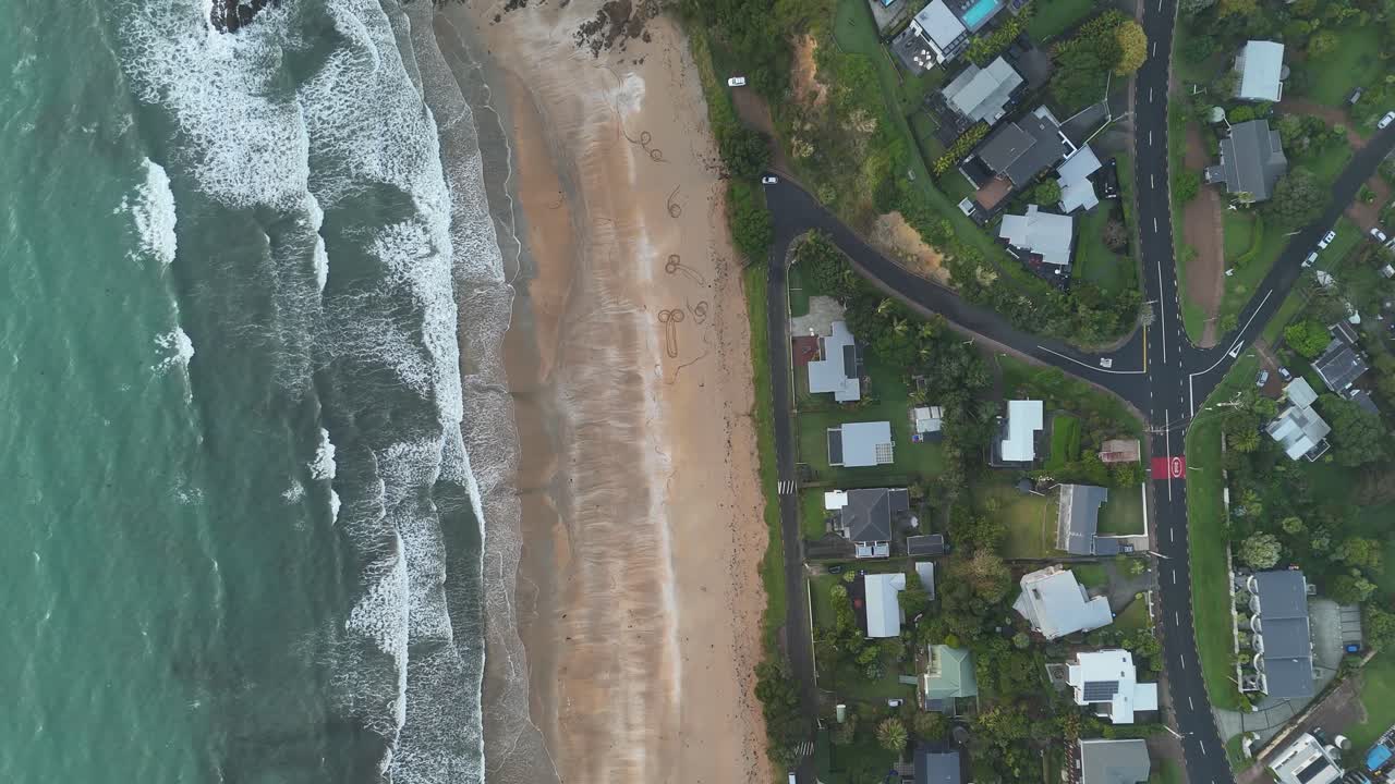 Waves and sandy beach of Zaiheke Island in New Zealand. Cloudy day with villas and mansion near beach. Aerial top down shot. Houses with ocean view.