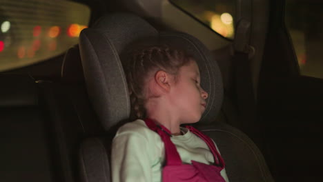 a close view of a child sleeping peacefully in a car seat, illuminated by the soft glow of ambient lights, with blurry view cars passing by outside