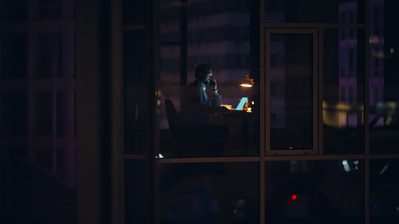 Direttore con vista sull'ufficio notturno seduto alla postazione di lavoro con il laptop. Lavoratore che fa tardi e che sta parlando